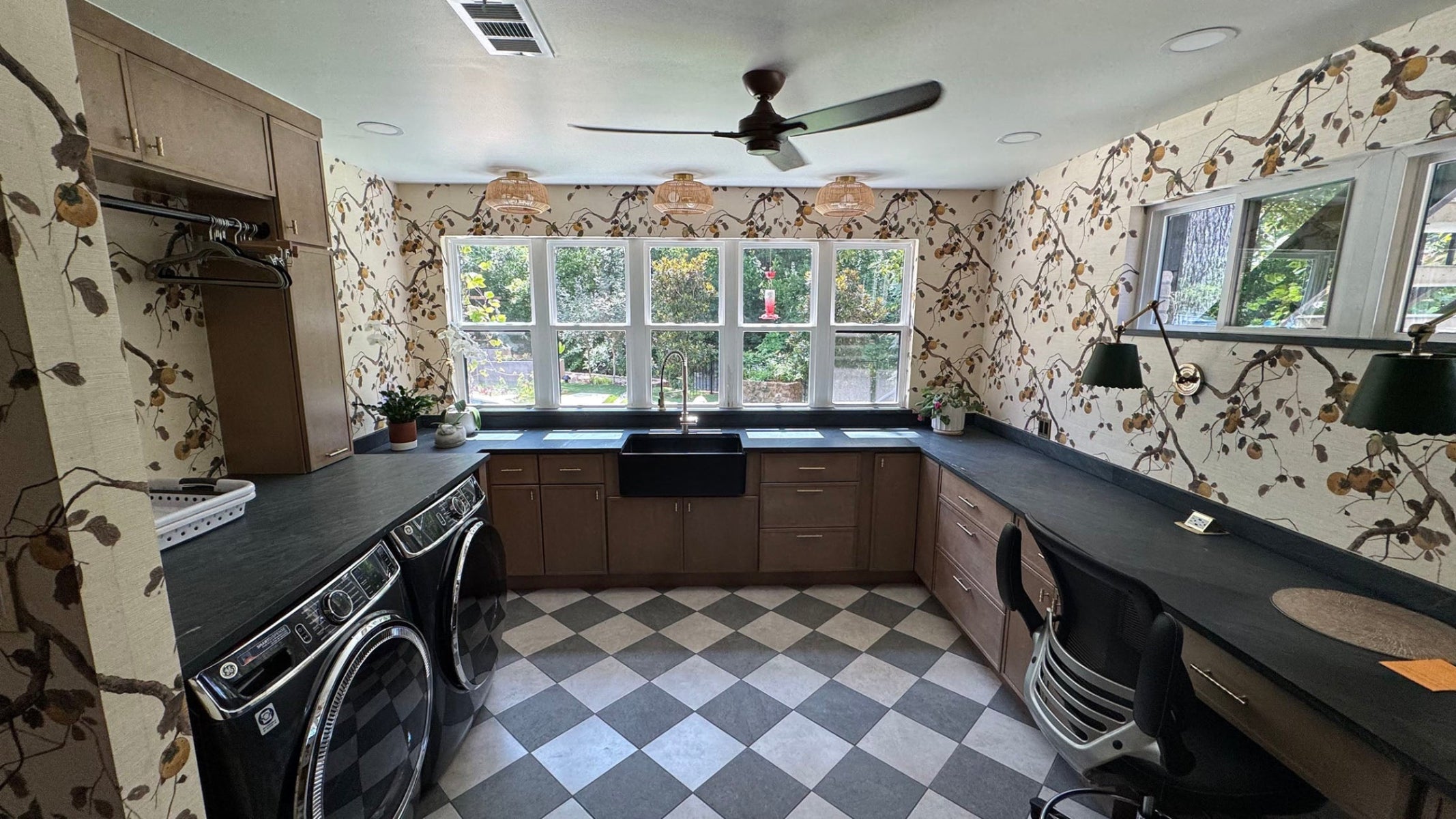 This bright and stylish multipurpose room combines a laundry area with a functional home office setup, featuring dark countertops, warm cabinetry, and bold floral wallpaper.